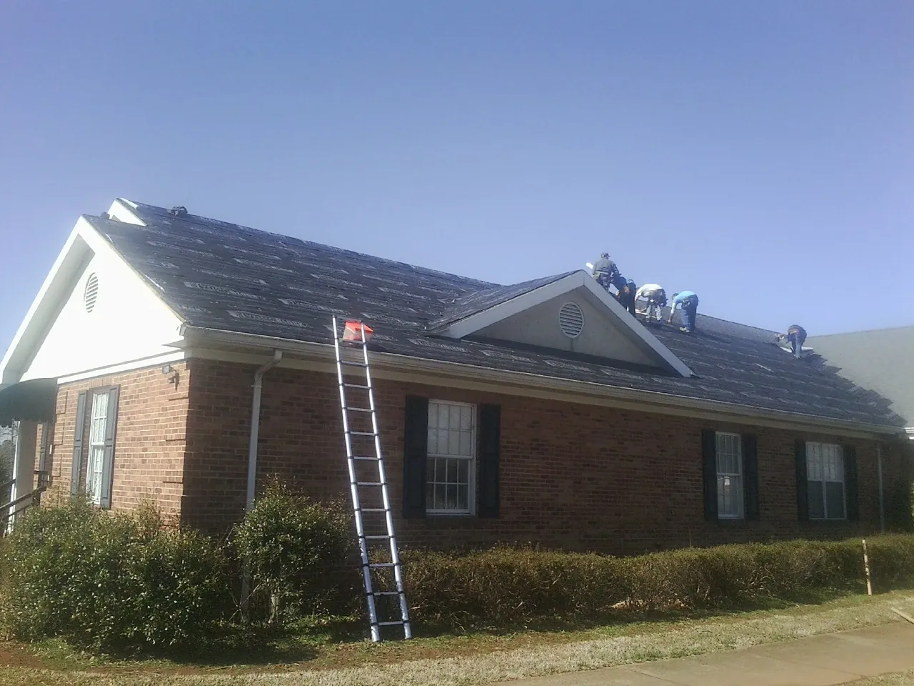Crew tearing shingles off a brick ranch roof with a ladder set — Emergency Roof Repair work by our crew in Mantachie