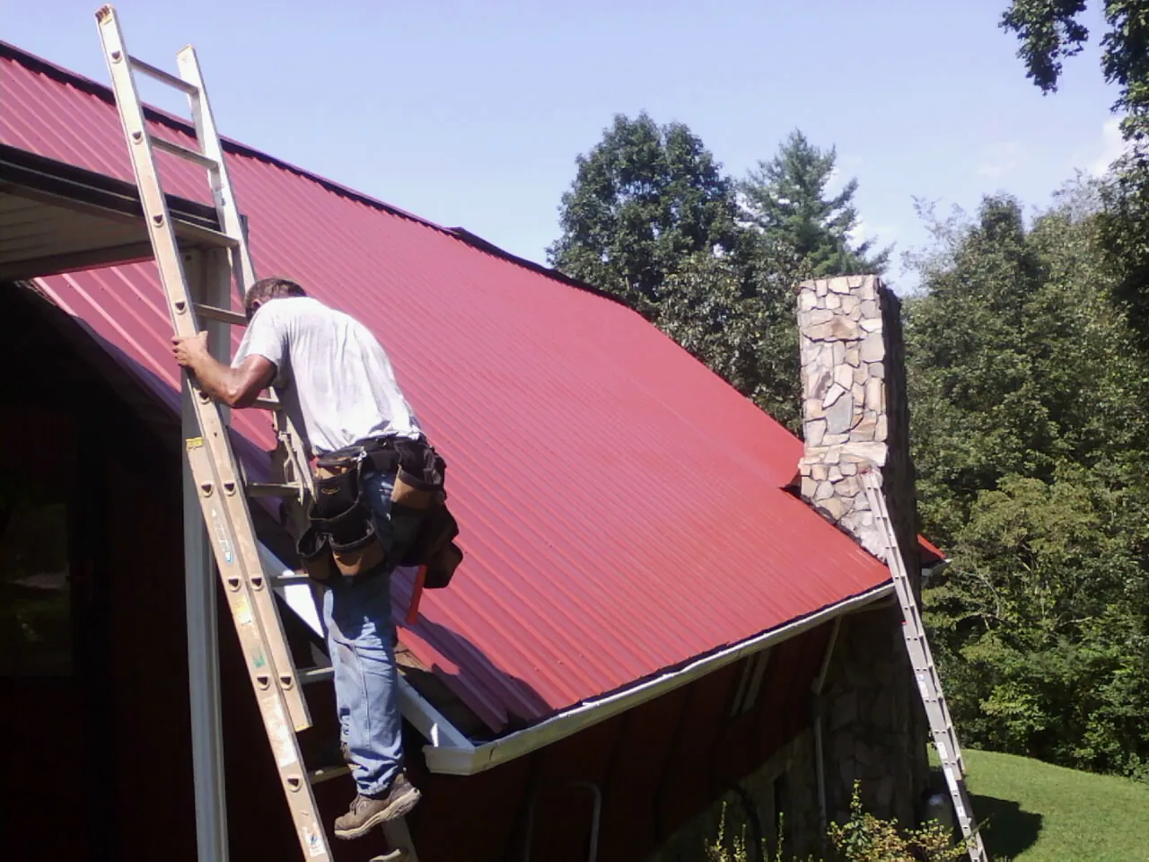 Roofer on a ladder beside a red metal roof and stone chimney — Soffit & Fascia Repair by Roofing Brothers in Mantachie